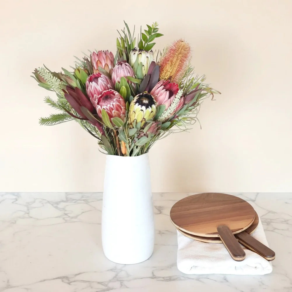 Bouquet of protea flowers in a white vase on a marble surface with wooden paddles.