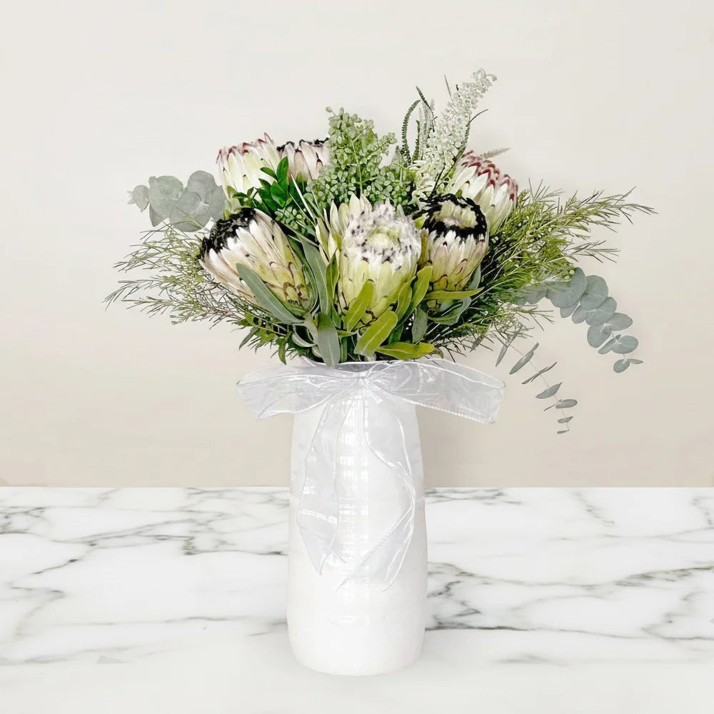 Protea arrangement in a white vase on a marble surface with a light background. Sympathy bouquet for special occasions.