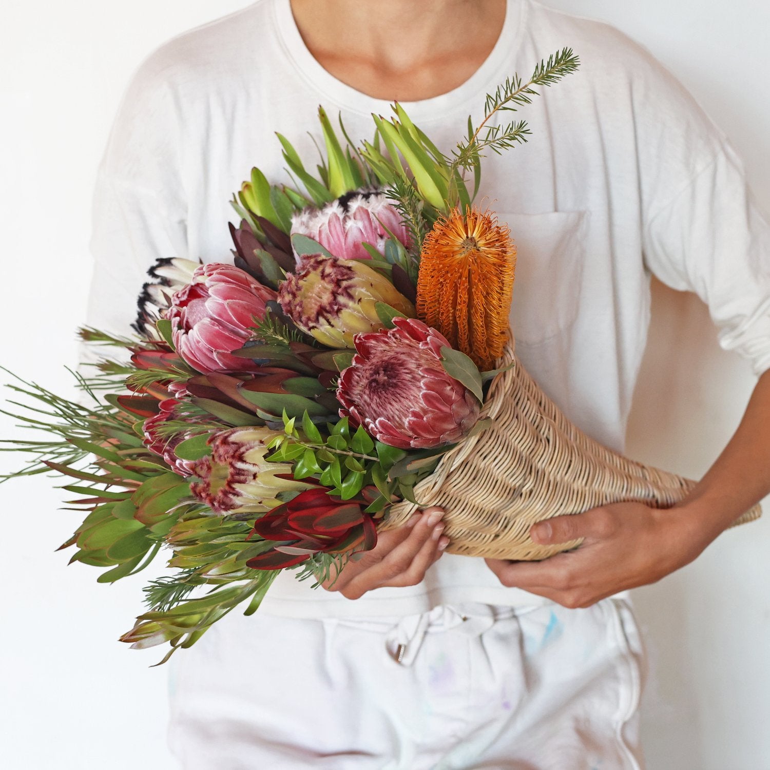 A person holding a colorful protea bouquet consisting of various protea and banksia blooms with green and red leucadendron foliage.
