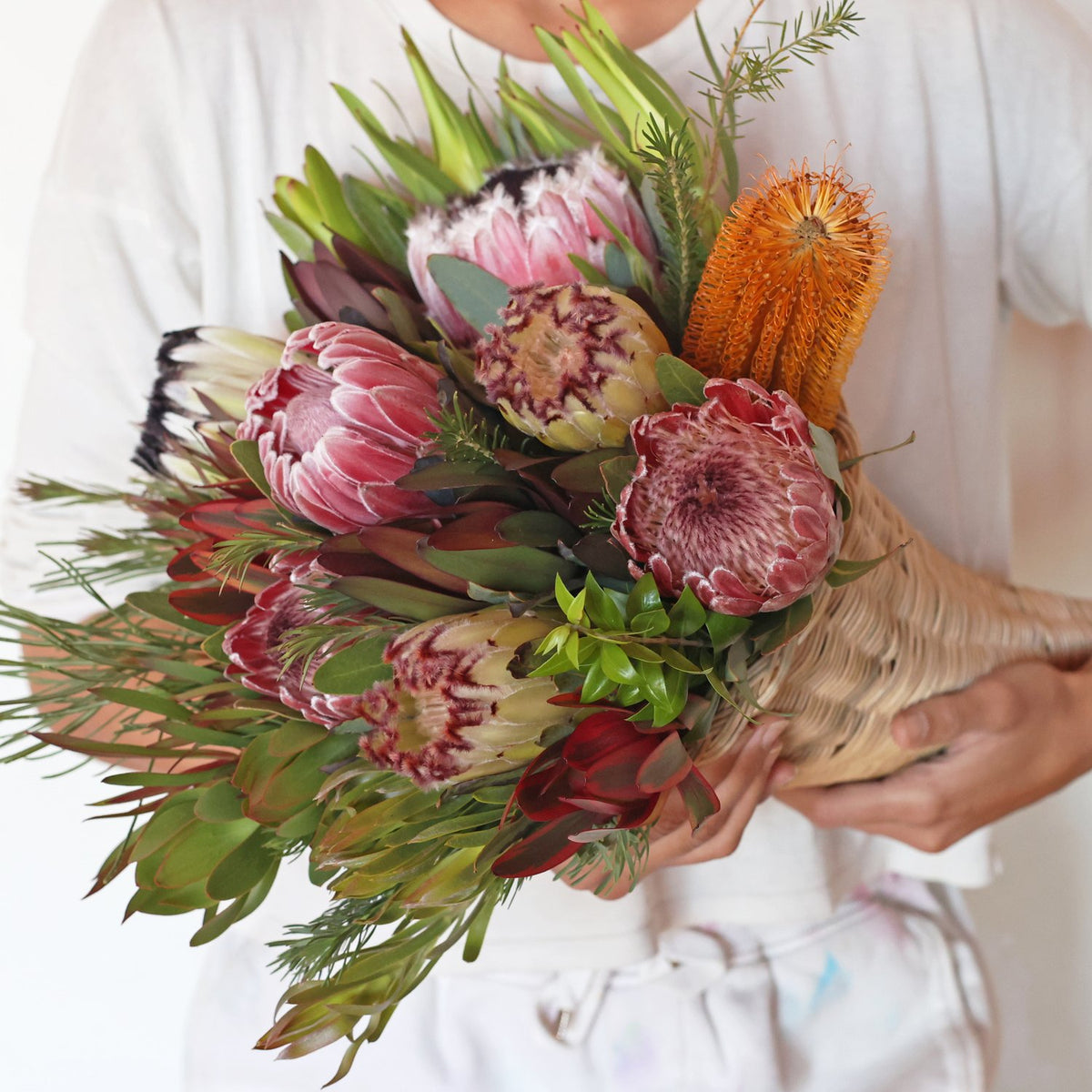 A person holding a colorful protea bouquet consisting of various protea and banksia blooms with green and red leucadendron foliage. Thanksgiving bouquet.